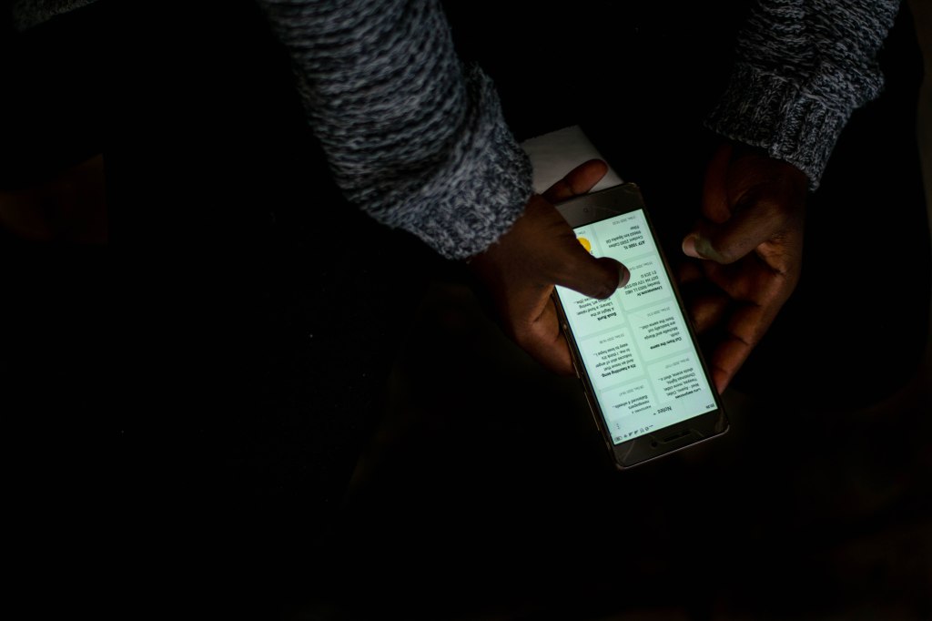 An upward shot of hands scrolling through notes on a phone screen in the dark. His arms are covered by a grey woolen cardigan.